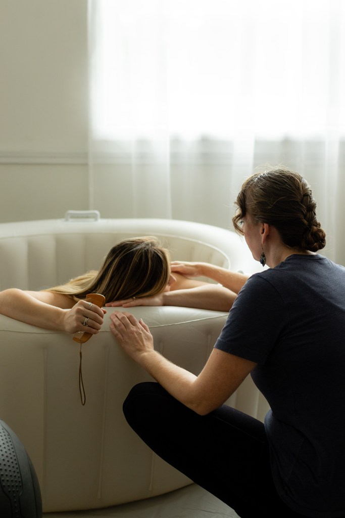A mother labors in a birthing pool, supported by her Doula, Ronae Cleland, in a calm, softly lit home birth setting in a birthing pool.